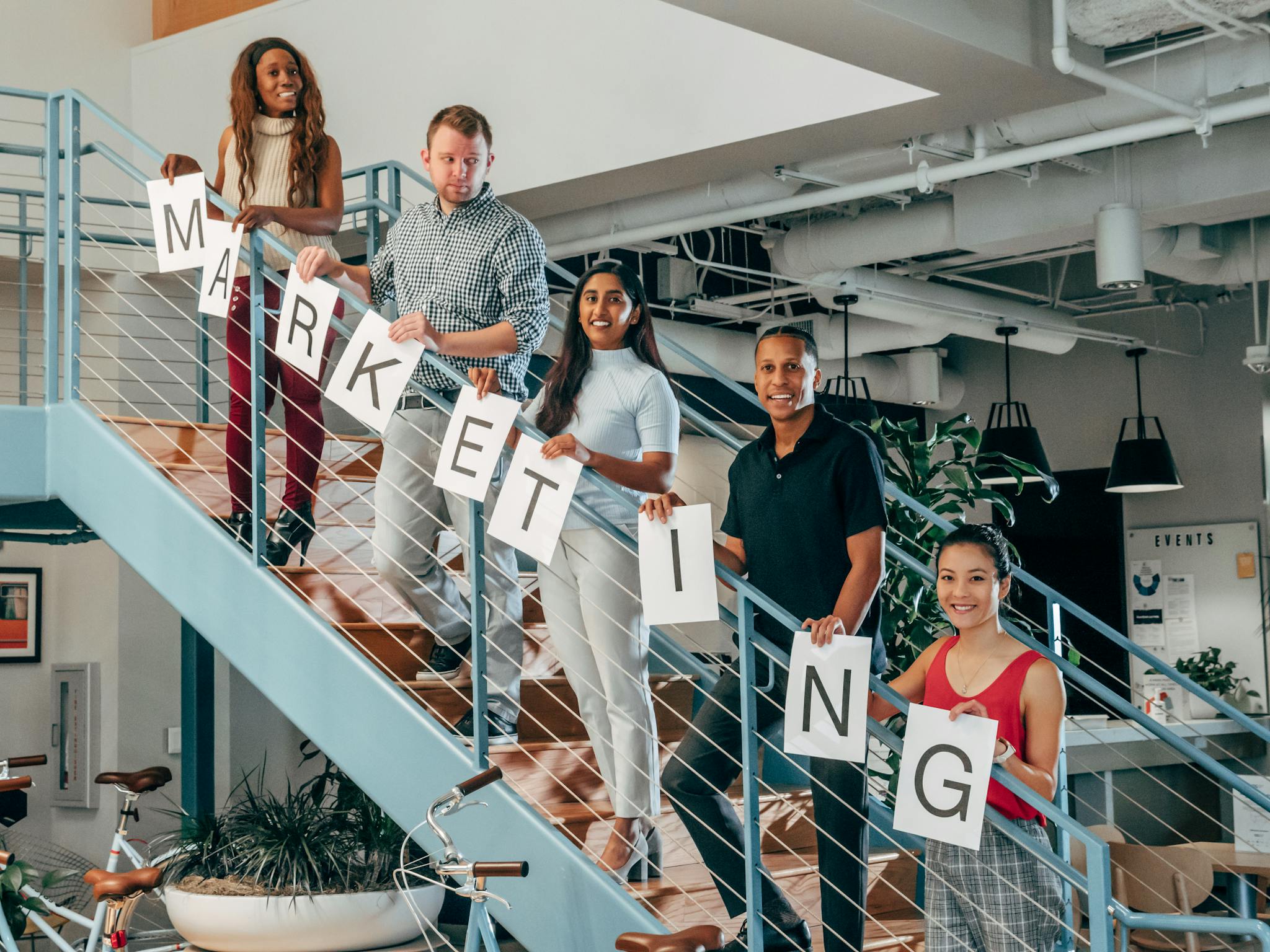 A diverse group of professionals holding 'marketing' signs on an office staircase.